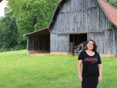 Woman standing on grass before a weathered wooden barn, wearing black shirt reading "North Carolina"