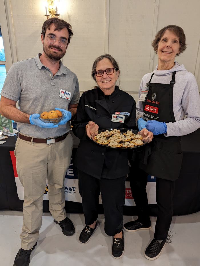 Three volunteers with NC State badges and blue gloves holding bowls and a tray of baked goods