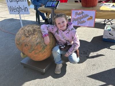 Girl kneels by giant pumpkin at "Pumpkin Weigh-in" station; sign reads "Free Pumpkin Pies"