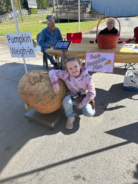 Girl kneels by giant pumpkin at "Pumpkin Weigh-in" station; sign reads "Free Pumpkin Pies"