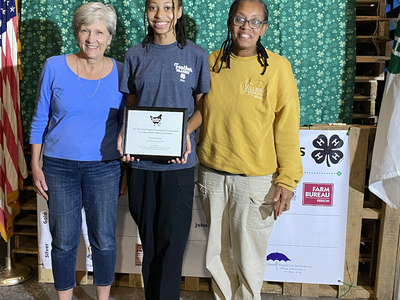 Three people standing before a green patterned backdrop; center person holds a framed certificate.