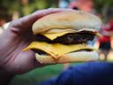A hand holds a hamburger. With a lawn and people gathering in the background.