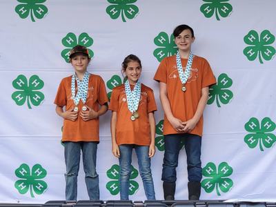 Three children in orange shirts wearing medals, standing in front of 4-H clover backdrop