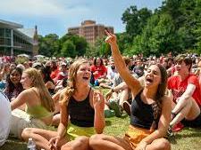 Two women sitting on grass cheering and pointing amid a large outdoor crowd