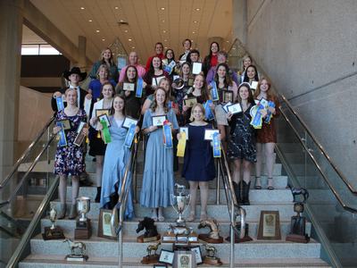 Group of students on staircase holding award ribbons and plaques, trophies displayed on steps