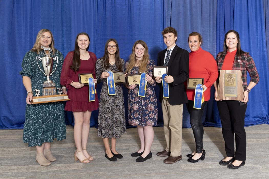 Seven people standing in front of a blue curtain holding trophies, plaques, and blue ribbons