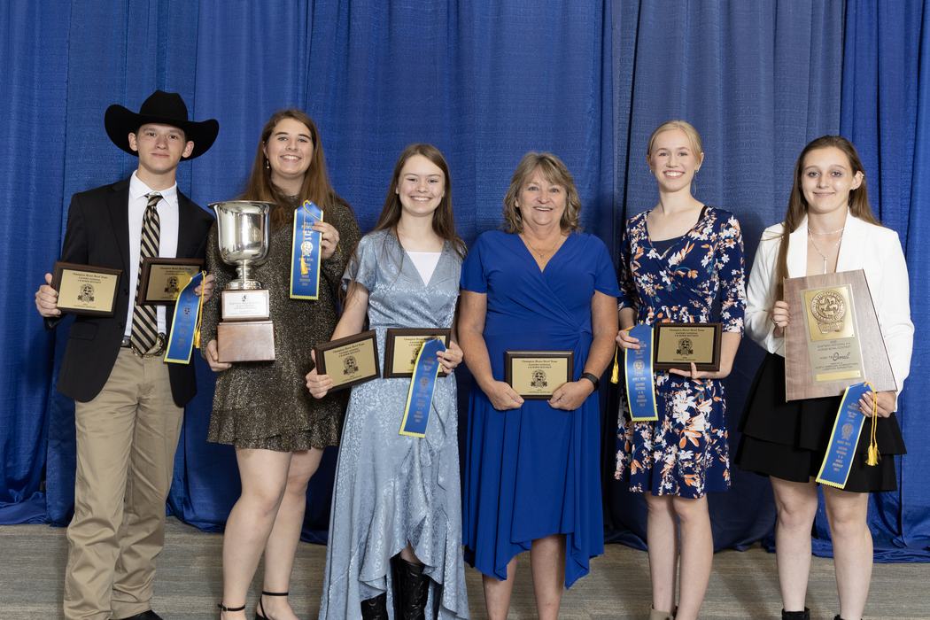 Six award recipients standing with trophies, plaques and blue ribbons in front of blue curtain