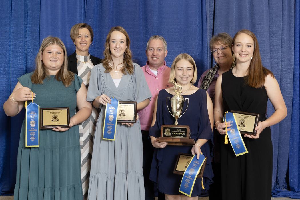 Seven people standing holding plaques, blue ribbons, and a trophy labeled "Team Communications Champion"