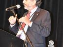 Bearded man in suit speaking into a microphone at a podium, crystal trophy on table nearby