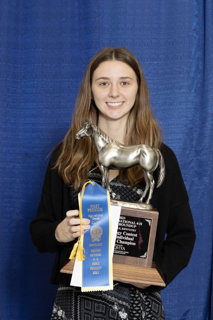 Woman holding horse trophy with blue ribbon reading "FIRST PREMIUM" and "Hippology Eastern National 4‑H 2023"
