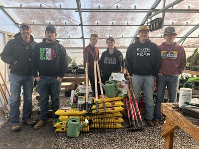 Six people in a greenhouse standing behind stacked potting mix, tools, and watering cans