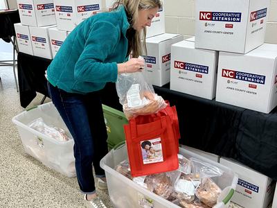 Woman placing packaged food into red bag beside stacked boxes labeled "NC Cooperative Extension"