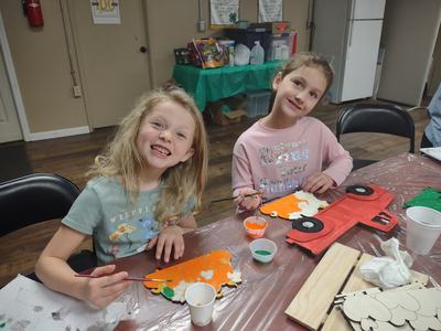 Two girls painting wooden crafts at a table; left girl's shirt reads "WILDFLOWER".