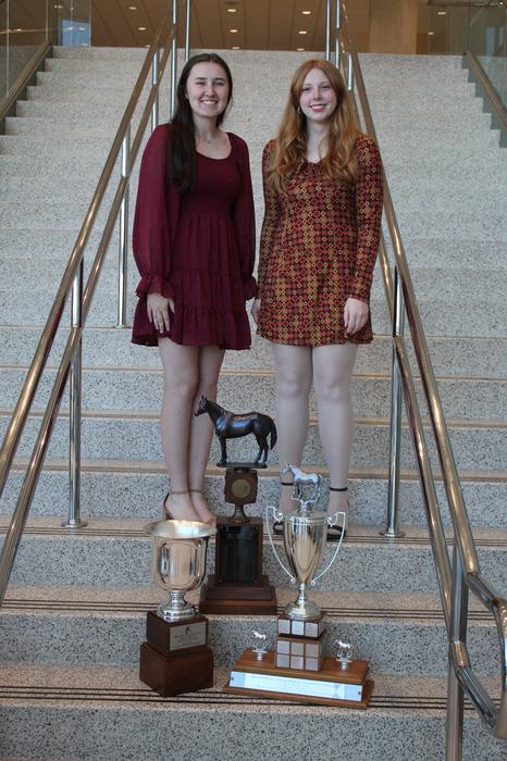 Two women standing on stairs behind three large trophies, two topped with horse figurines.