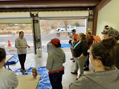 Woman speaking to a group inside a garage-style room with open doors and parked cars