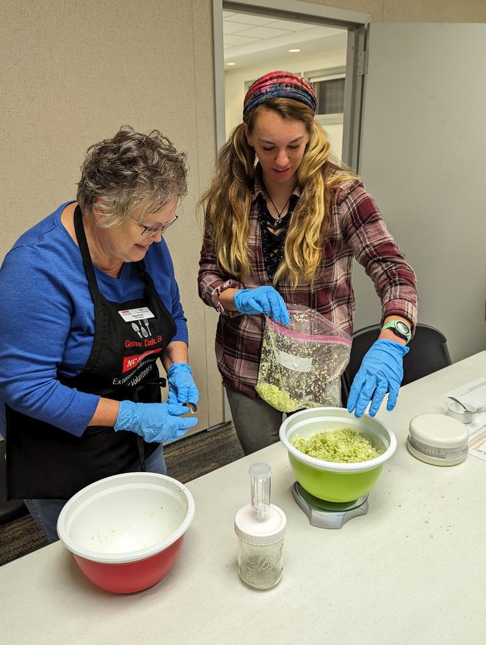 Two people wearing gloves transfer chopped green vegetables from a plastic bag into a green bowl