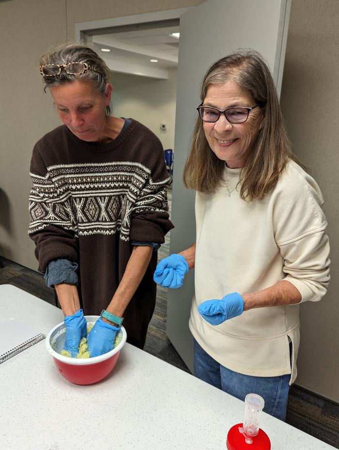 Two women wearing blue gloves, one mixing contents in a red bowl on a table