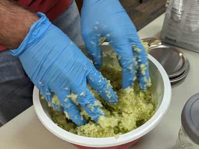 Hands in blue gloves mixing shredded green cabbage in a white-and-red bowl
