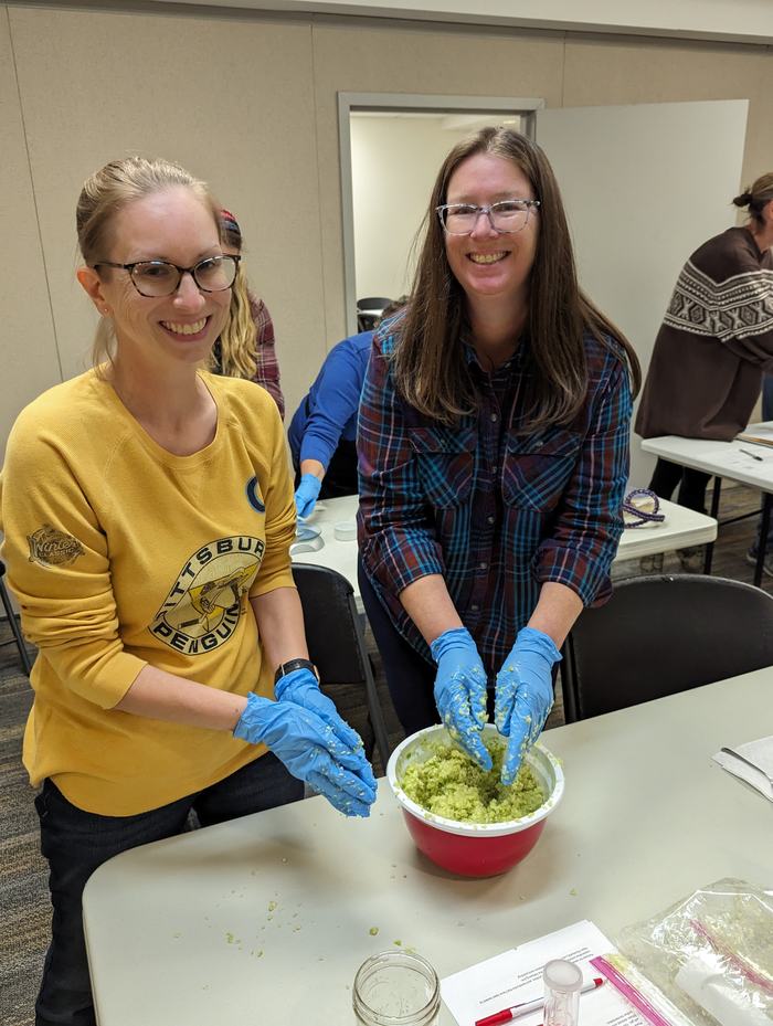 Two women wearing blue gloves mixing mashed green food in a red bowl; left shirt reads "PITTSBURGH PENGUINS"