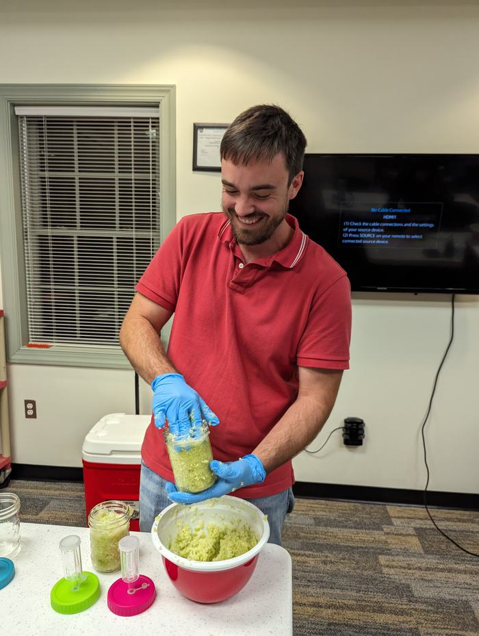 Person in red shirt wearing blue gloves packing green mashed food into a jar over a bowl