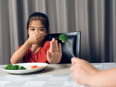 Child covering mouth and holding a hand out to refuse broccoli being held by an adult.