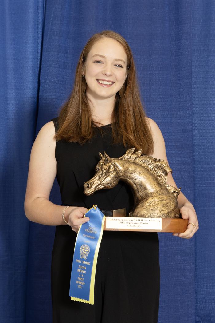 Young woman holding a horse-head trophy and a blue ribbon reading "FIRST PREMIUM" and "Public Speaking"