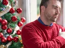Man in red sweater with arms crossed standing by window beside decorated Christmas tree