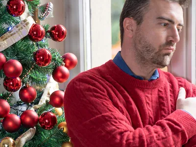 Man in red sweater with arms crossed standing by window beside decorated Christmas tree