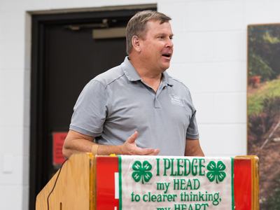 Man speaking at a podium with banner reading "I PLEDGE my HEAD to clearer thinking, my HEART"