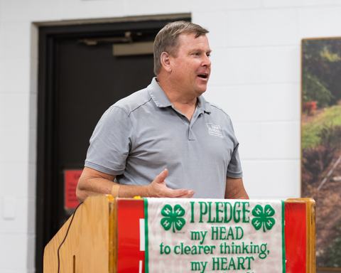 Man speaking at a podium with banner reading "I PLEDGE my HEAD to clearer thinking, my HEART"