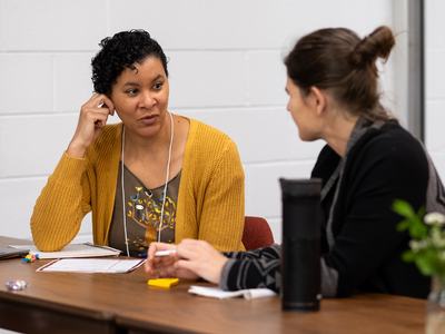 Two people seated at a table conversing with papers, a notebook, and a bottle.