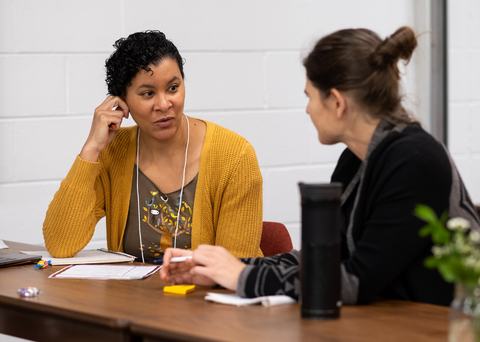 Two people seated at a table conversing with papers, a notebook, and a bottle.