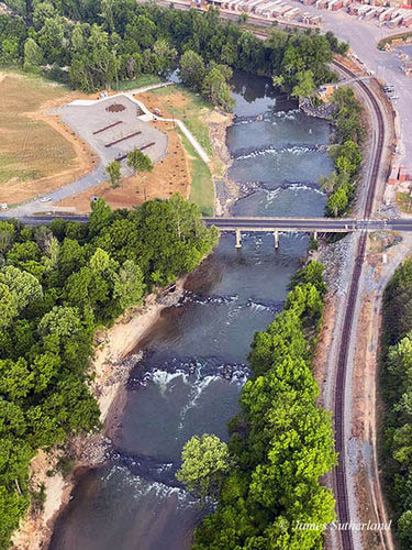 Aerial view of Madison River Park with new water fall systems in place of the dam at the bridge. Shows six small waterfalls along the Dan River lined with trees, beaches and a railroad track on one side. The bridge runs across the river in the middle the six waterfalls pictured. 
