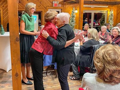 Two women embracing at a crowded indoor dinner event as nearby guests applaud