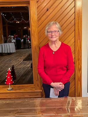 Older woman in red sweater standing indoors by wood-paneled wall and window.