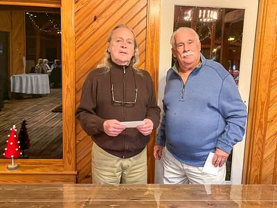Two older men standing indoors by a wood-paneled wall, each holding a white envelope.