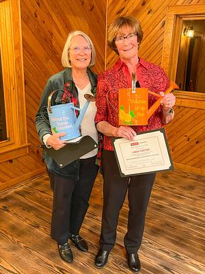 Two women holding awards: blue sign "Behind the Scenes Volunteer" and orange plaque "Volunteer of the Year"