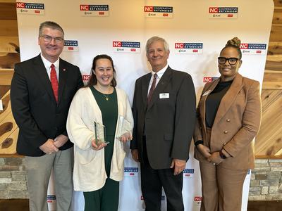 Four people standing at NC Cooperative Extension backdrop; second person holds two glass awards.