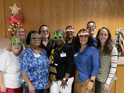 Group of nine people wearing festive novelty glasses posing in a room with small decorated Christmas trees