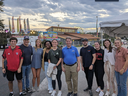 Group of students posing with director of fair in front of fairgrounds building and ferris wheel in the background.