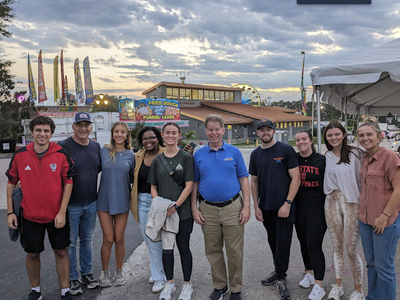 Group of students posing with director of fair in front of fairgrounds building and ferris wheel in the background.