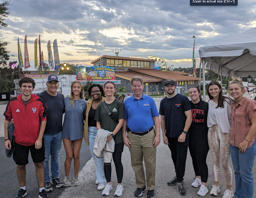 Group of students posing with director of fair in front of fairgrounds building and ferris wheel in the background.