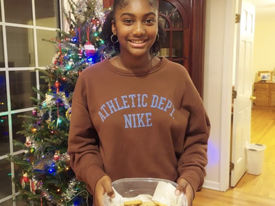 Teen holding container of cookies by a decorated Christmas tree; sweatshirt reads "ATHLETIC DEPT NIKE"