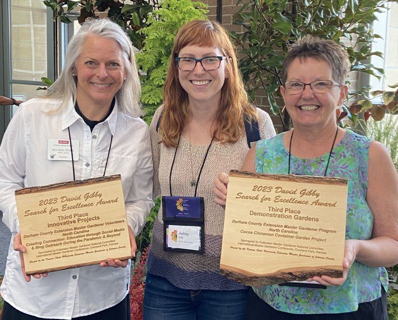Three women smile as they display wooden plaques inscribed with the heading 2023 David Gibby Search for Excellence Award.