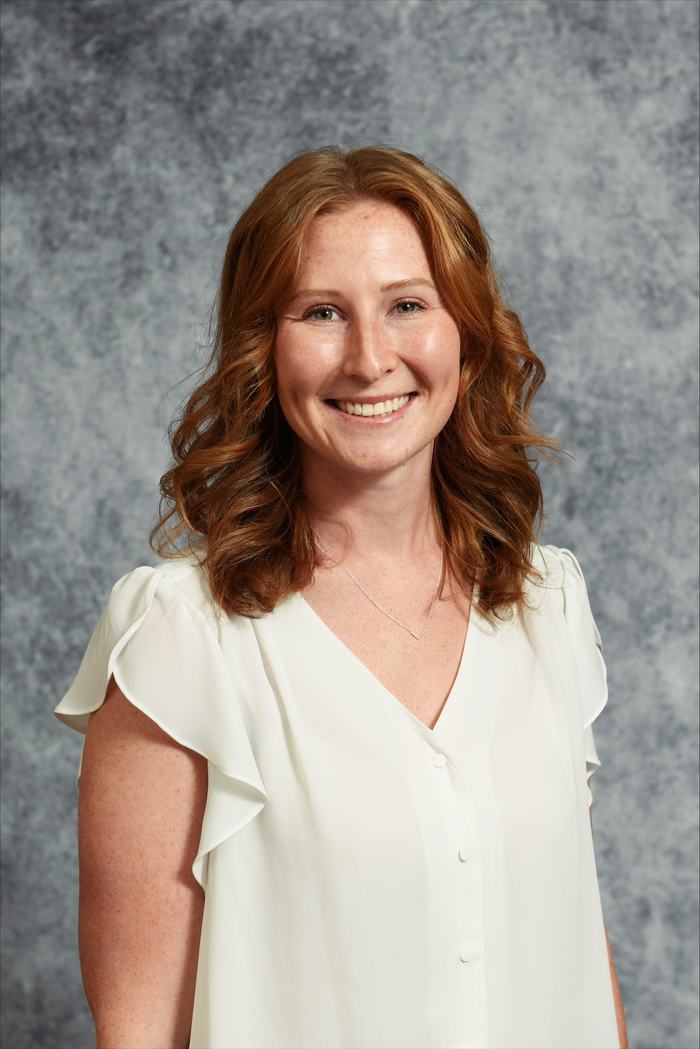 Woman with red hair, white blouse and light necklace against gray mottled backdrop