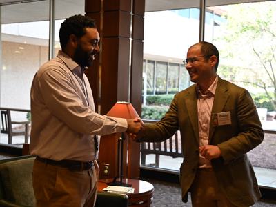 Two men in business attire shaking hands in a lobby near a window.