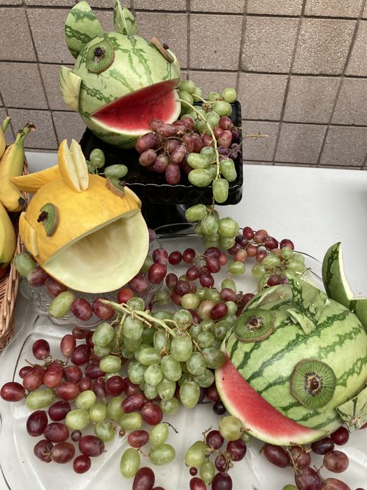 Three melons cut and decorated to look like fish sit amongst a platter of fruit on a white table.