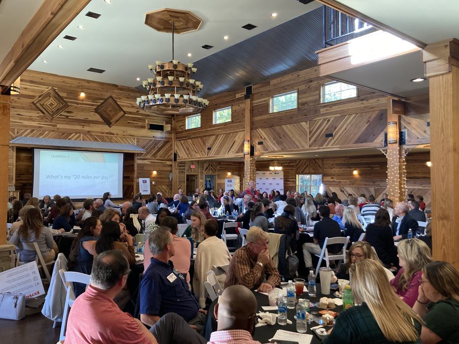 A barn event space full of people seated at round tables, listening to the keynote speaker present at the front of the room.