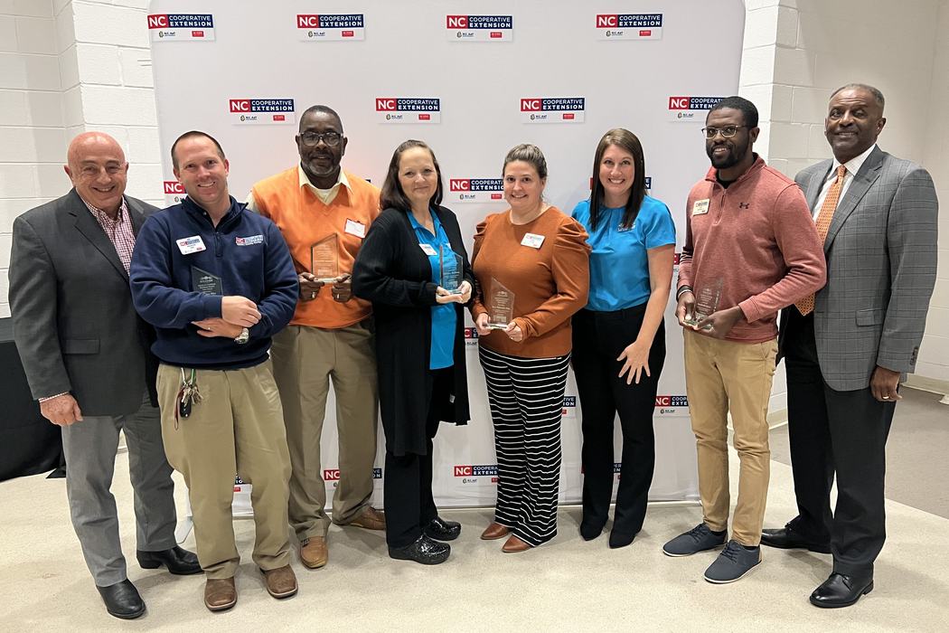 A group of Extension professionals stand in front of a N.C. Cooperative Extension step-and-repeat banner. Each member of the award winning team has a glass plaque in their hands.