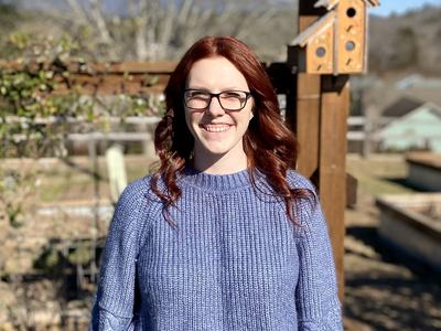 Person with red hair and glasses wearing blue sweater standing by a wooden trellis with birdhouse